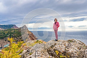 Teen tourist girl standing on mountain cliff against beautiful seascape in cloudy day. Young lady posing on background of sea,