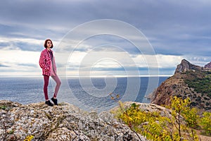 Teen tourist girl standing on mountain cliff against beautiful seascape in cloudy day. Young lady posing on background of sea,