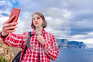 Teen tourist girl taking selfie with smartphone against beautiful seascape in cloudy day. Young lady posing on background of sea,