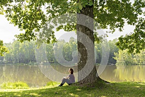 Teen young girl woman sitting on grass under huge high big old oak,trunk tree near lake,river, meditating,relaxing and smiling in