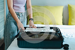 A teenage Asian woman with long hair sits on a bed in her bedroom, folding clothes into a large suitcase to prepare for weekend