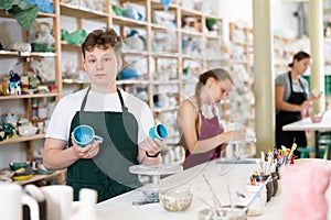 Teenage boy in an apron demonstrates clay mugs, which he himself painted with colored paints