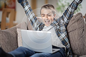 Teenage boy with arms raised up celebrating success using laptop on sofa at home