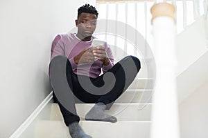 Teenage Boy Being Bullied By Text Message Sitting On Stairs At Home