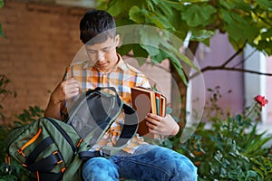 A teenage boy, being a student, is sitting on a bench near the school building, he takes a book or other school supplies out of