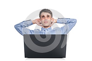 A teenage boy in a blue shirt sits in front of a laptop with his hands behind his head and looks up, thinking. Isolated