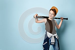 Teenage boy in cap, undershirt, jeans and hoodie tied around his waist. Holding black baseball bat, posing on blue background