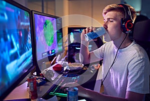 Teenage Boy Drinking Caffeine Energy Drink Gaming At Home Using Dual Computer Screens At Night