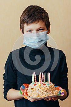 Teenage boy in face mask holds cake with candles in hands and celebrates birthday isolated without friends. Sad because of the lim