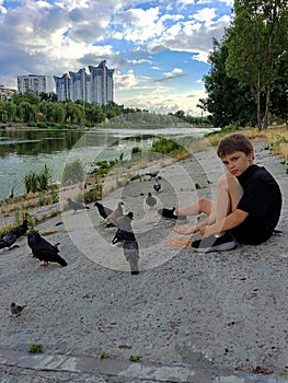 A teenage boy feeds pigeons from his hands on the river bank in the park