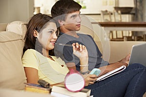 Teenage Boy And Girl Sitting On Sofa At Home Doing Homework Using Laptop Computer Whilst Holding Mobile Phone