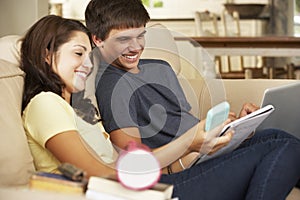 Teenage Boy And Girl Sitting On Sofa At Home Doing Homework Using Laptop Computer Whilst Holding Mobile Phone