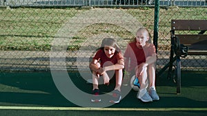 Teenage boy and girl in sports uniforms sit on football pitch near bench at sunlight