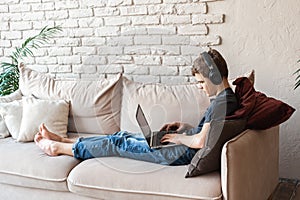 A teenage boy in headphones and with a laptop sits at home on the couch and communicates with peers using wireless