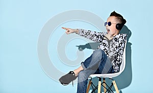 Teenage boy in headphones, sunglasses and casual clothes. Pointing at something, sitting on white chair against blue background