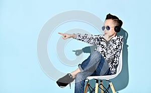 Teenage boy in headphones, sunglasses and casual clothes. Pointing at something, sitting on white chair against blue background