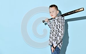 Teenage boy in hoodie and jeans. He is grinning, holding black shabby baseball bat or club, posing sideways on blue background