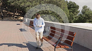 teenage boy in jeans and a shirt walks through the park with phone in his hands and sits on a bench