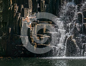 Teenage boy jumping in waterfall lake. Falling water streams flow on black volcanic stone cascades. Rochester Falls waterfall -