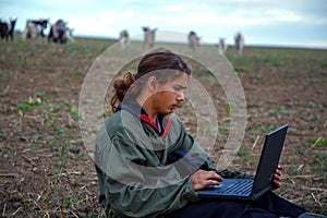 A teenage boy with a laptop grazes goats in a field. A goat herder in a field with a laptop communicates over the Internet.
