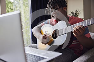 Teenage Boy Learning To Play Acoustic Guitar With Online Lesson On Laptop Computer At Home