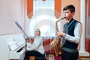 A teenage boy learns to play the saxophone in a music lesson to the accompaniment of a female teacher on the piano