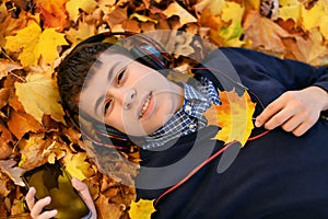 A teenage boy listens to music with headphones, lies on yellow autumn maple leaves, a bright sunny day, beautiful nature in autumn