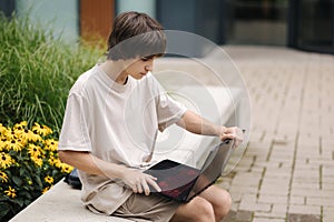 Teenage boy open his laptop and try to study outdoors. Sitting on a bench under a house in a residential area