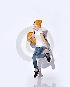 Teenage boy with orange travel bag, in hat, t-shirt, blue jeans, black sneakers. He jumping up, smiling, posing isolated on white