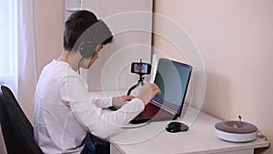 Teenage boy playing game on his pc computer in white room. Gamer capture video on web camera on laptop and phone on