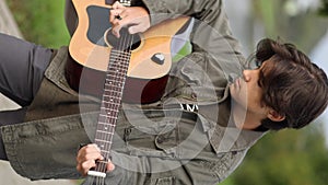 Teenage boy playing guitar outdoor in park. Long hair boy alone play on guitar. Autumn mood. Vertical video