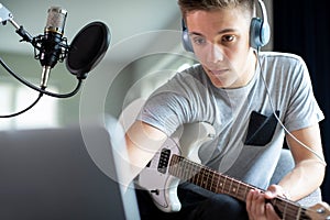 Teenage Boy Playing Guitar And Recording Music Onto Laptop At Home