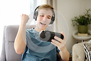 Teenage boy is playing a portable game console. Guy sits in a chair in the living room and plays a gadget. Gambling young person