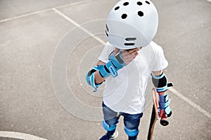 Teenage boy in protective helmet and gears holding a skateboard, looking down and wiping tears from his eyes