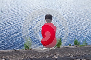 A teenage boy in a red hoodie is squatting on the shore of a bay sea or river