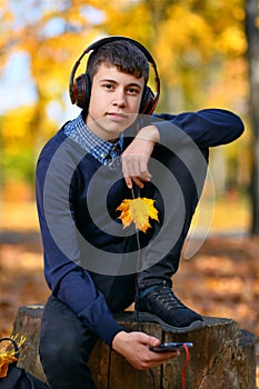 A teenage boy sits in autumn park and listens to music with headphones, trees with yellow leaves on a bright sunny day