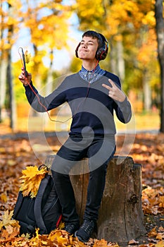 A teenage boy sits in autumn park and listens to music with headphones, trees with yellow leaves on a bright sunny day