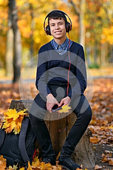 A teenage boy sits in autumn park and listens to music with headphones, trees with yellow leaves on a bright sunny day