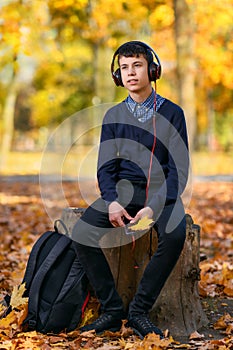 A teenage boy sits in autumn park and listens to music with headphones, trees with yellow leaves on a bright sunny day