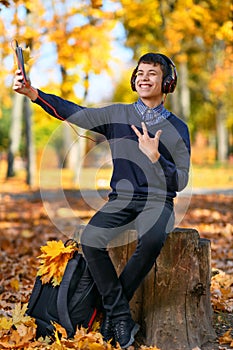A teenage boy sits in autumn park and listens to music with headphones, trees with yellow leaves on a bright sunny day