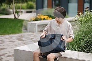 Teenage boy sits on bench and open his laptop bag