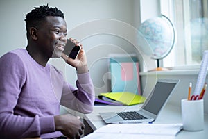 Teenage Boy Sitting At Desk Doing Homework Assignment On Laptop Talking On Mobile Phone