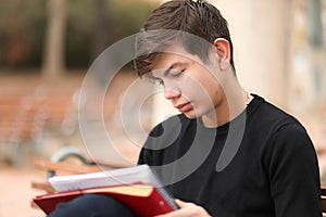Teenage boy is sitting in a park while doing school homework with a serious face and concentration