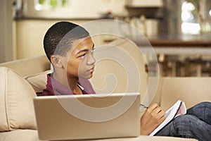 Teenage Boy Sitting On Sofa At Home Doing Homework Using Laptop Computer Whilst Watching TV