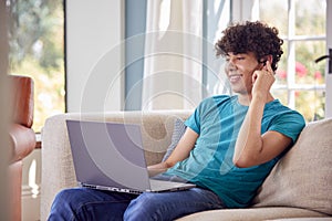 Teenage Boy Sitting On Sofa At Home Using Laptop Computer And Talking On Mobile Phone