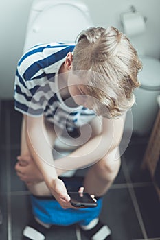 Teenage boy sitting on a toilet with his mobile