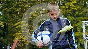 Smiling teenage boy with a soccer ball in his hand and soccer boots on the shoulder against the background of the