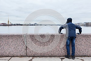 Teenage boy standing on granite embankment and looking at Peter and Paul Fortress across Neva river, Saint-Petersburg, Russia