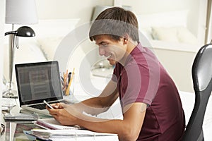 Teenage Boy Studying At Desk In Bedroom Using Mobile Phone