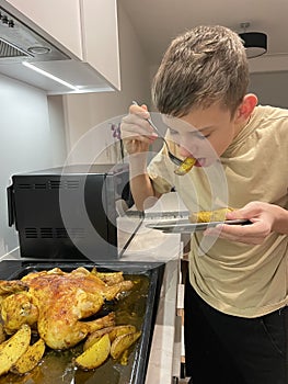 A teenage boy tastes baked chicken and potatoes in the kitchen.